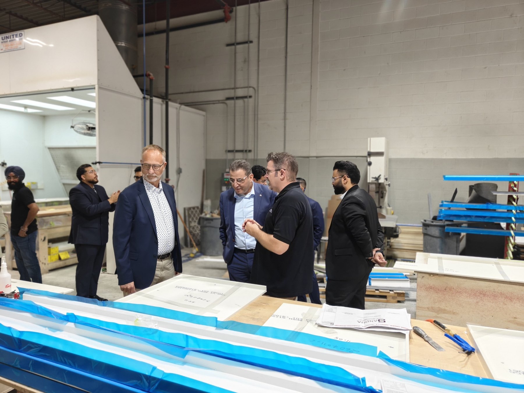 Members of Parliament and guests gather around a workbench with protected finished panels and Sandha production drawings.