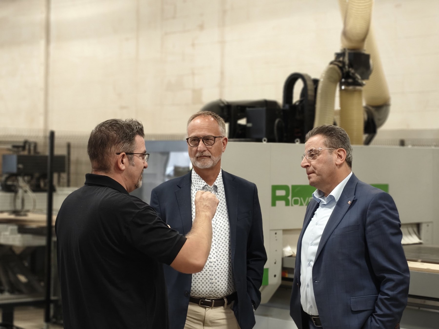 A Sandha Woodworks representative speaks with two Members of Parliament in front of a CNC machining centre.