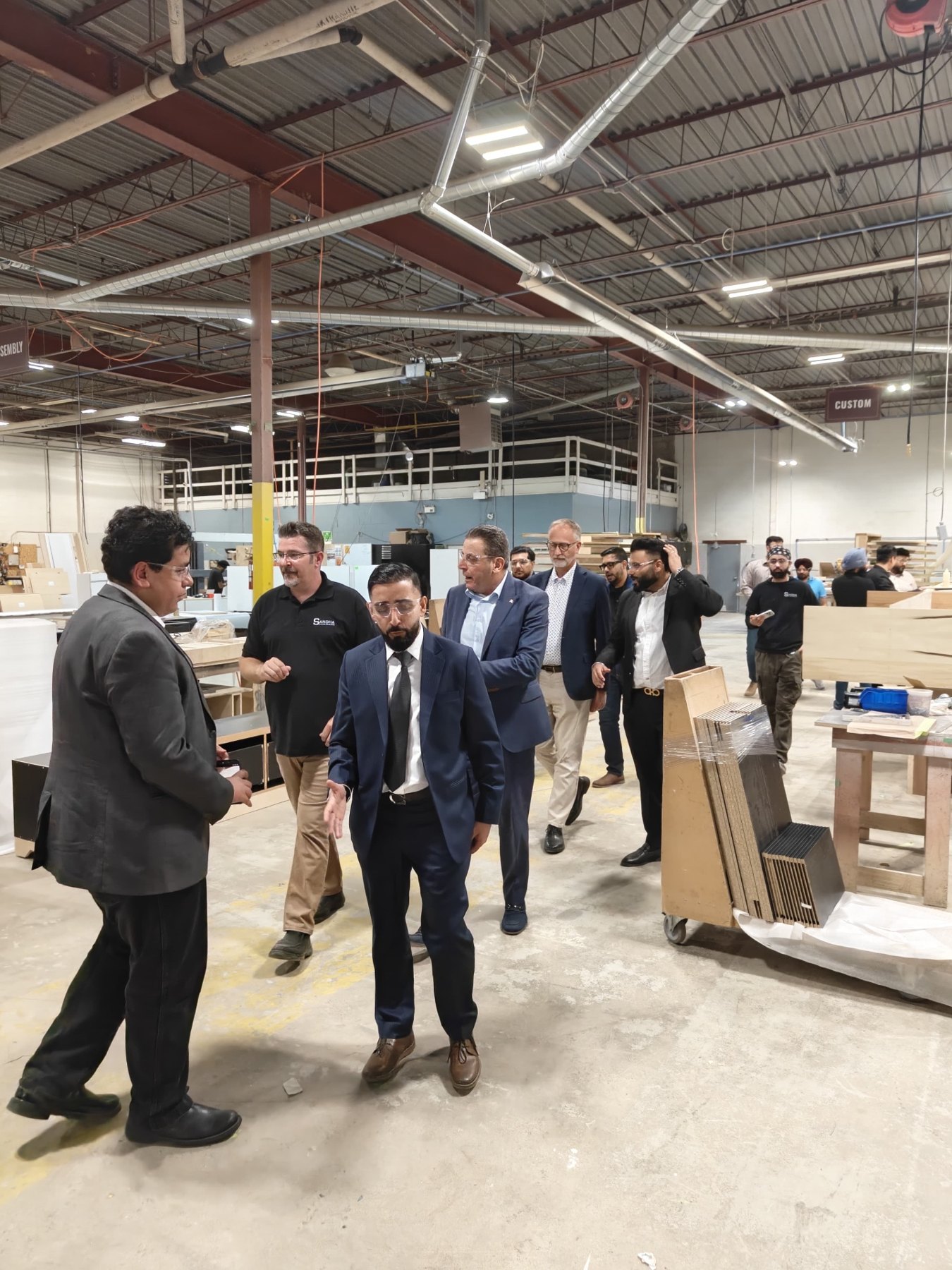 Visiting delegation walks through the Sandha Woodworks production floor with company representatives, assembly and custom areas visible overhead.